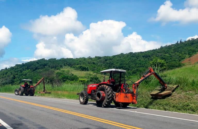 Dois tratores realizando roçagem na vegetação ao longo da rodovia BA-052, em um dia ensolarado.